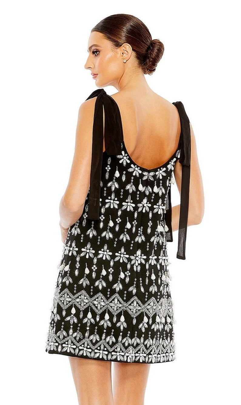 Woman wearing the Short Wedding Guest Dress 10937 by Mac Duggal—a sleeveless black dress with white beaded floral patterns and black ribbon straps—shown from the back against a white background.